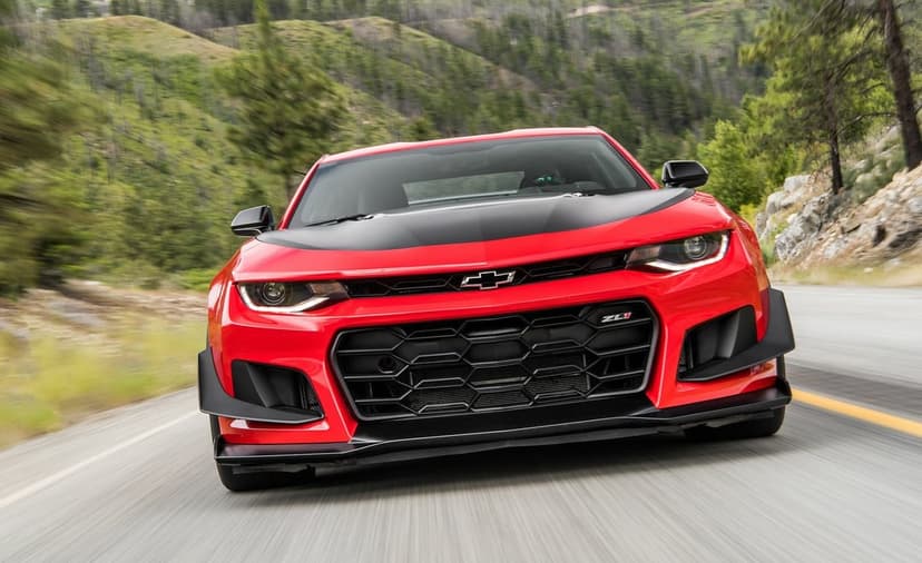 Red Chevrolet Camaro ZL1 with a black hood driving fast on a mountain road with blurred green trees.