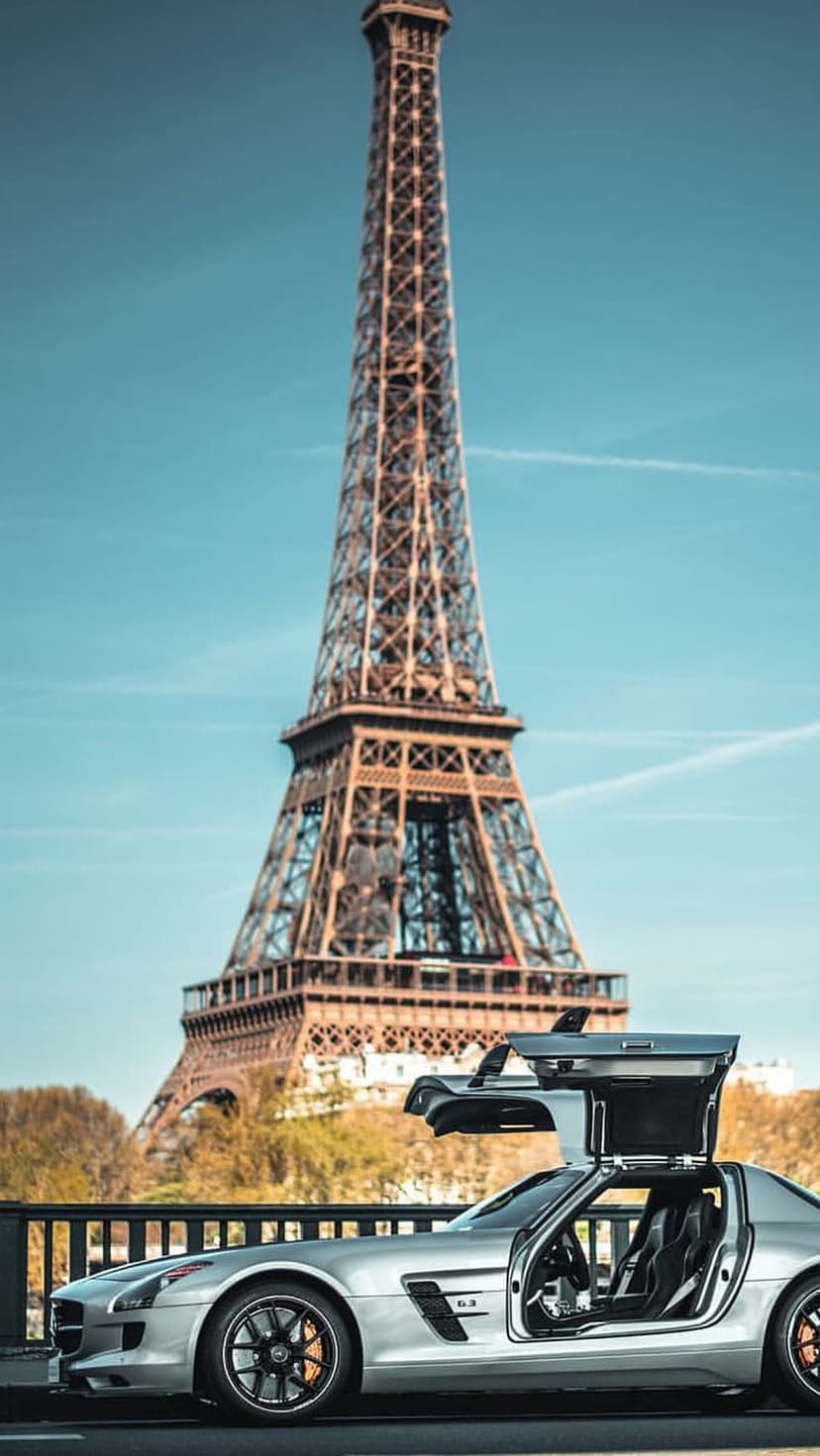 Silver Mercedes SLS AMG with open gullwing doors parked on a Paris bridge with the Eiffel Tower in the background.