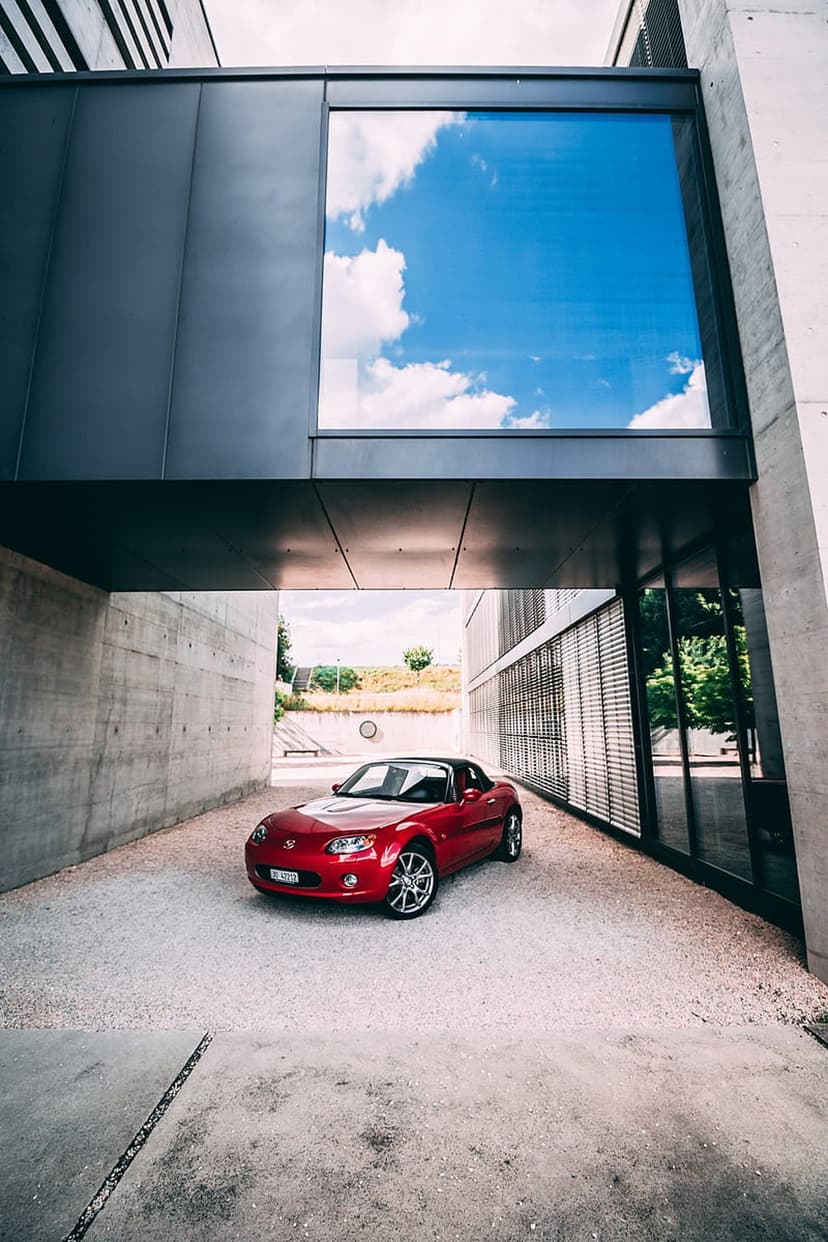 Red convertible roadster parked under a concrete cantilevered roof with a reflection of blue sky in glass.