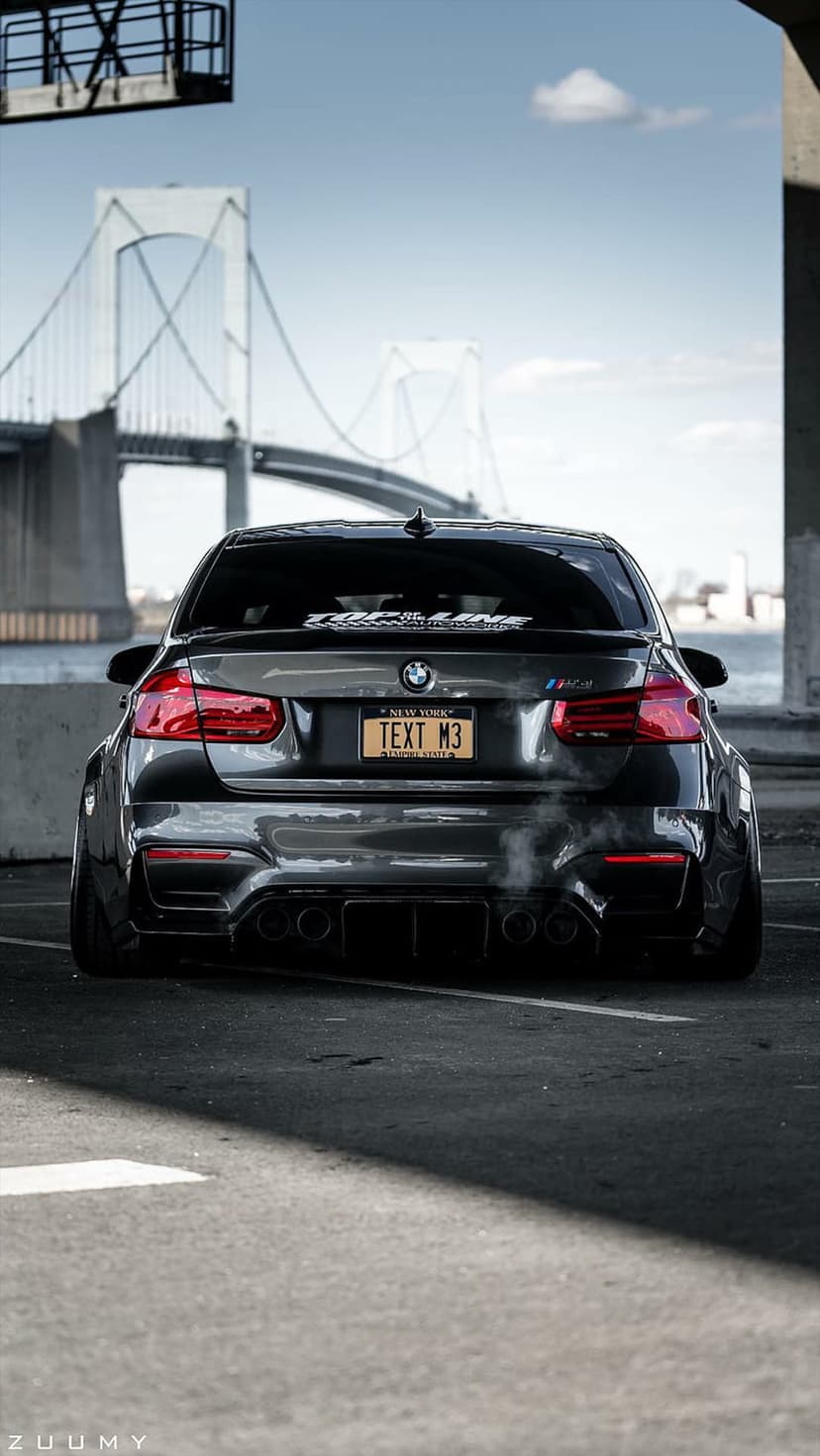 BMW M3 sports sedan rear view with exhaust smoke parked under a massive suspension bridge against a blue sky.