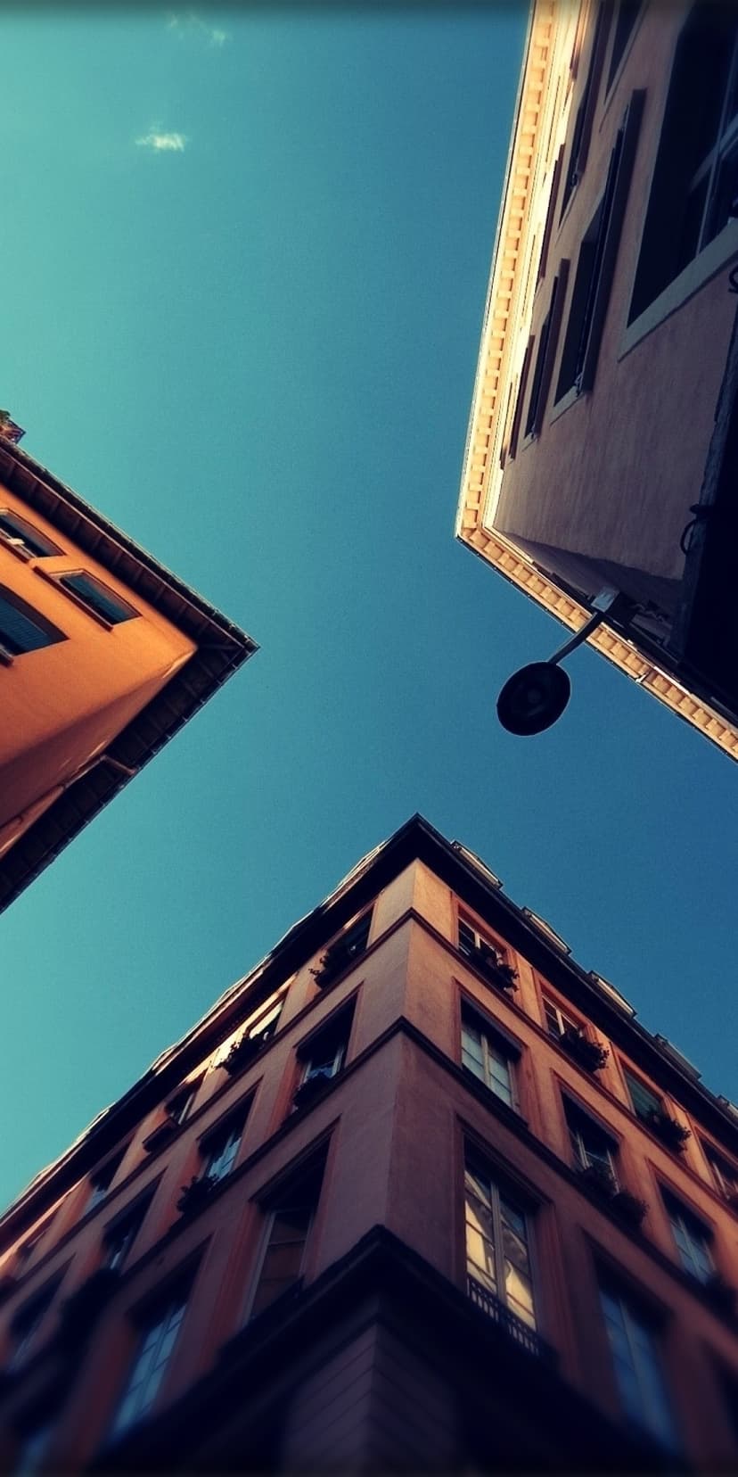 Three warm-toned buildings angle upward toward a clear blue sky with a silhouetted streetlamp in the foreground.