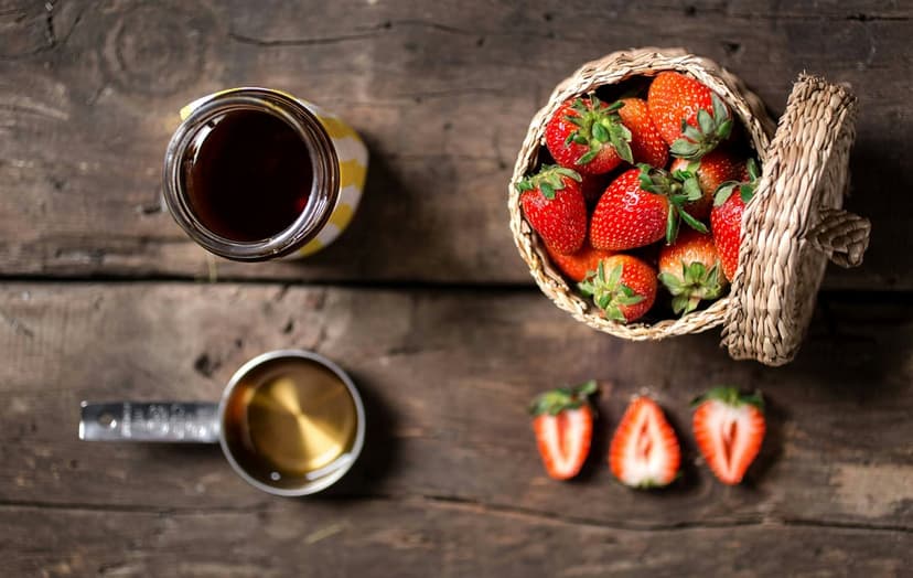Strawberries in a wicker basket beside a jam jar and metal measuring cup resting on a rustic wooden tabletop.