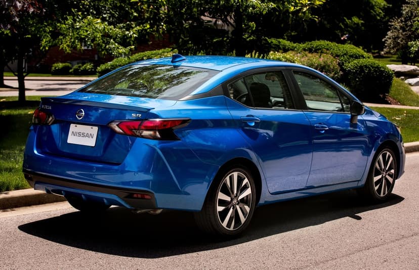 Rear three-quarter view of a metallic blue Nissan Versa SR parked on an asphalt street with green trees behind.