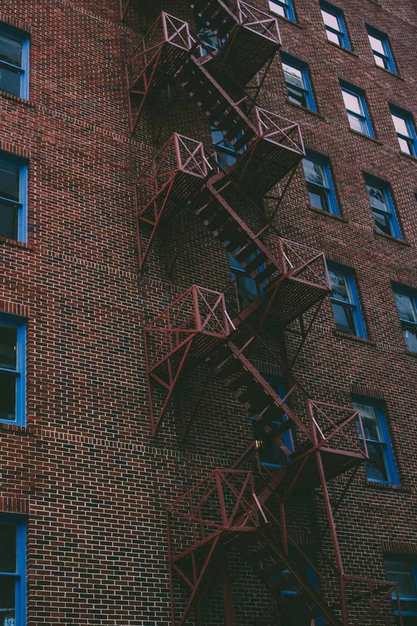 Red metal fire escape stairs attached to an old brick wall with vibrant blue window frames in an urban setting.