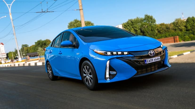Blue Toyota Prius hybrid car driving on an asphalt road with a clear sky and trees in the background.