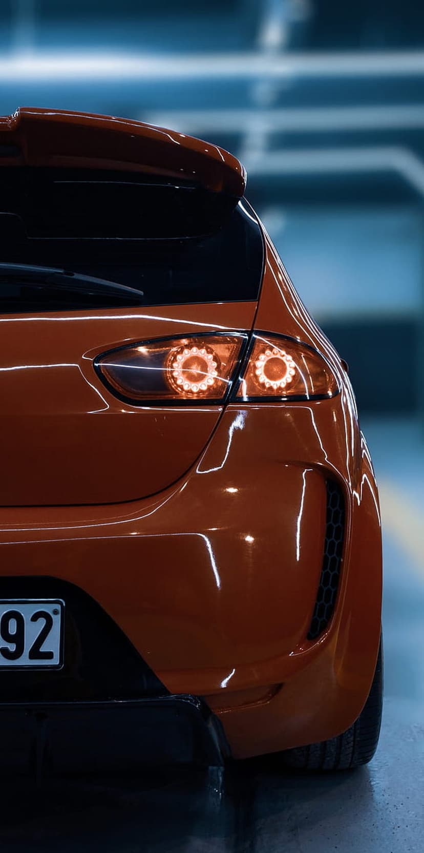 Orange sports car rear taillights and spoiler glowing with warm light on a glossy metallic vehicle surface.