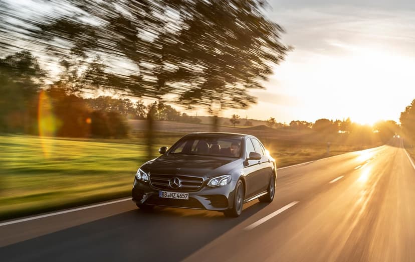 Dark gray Mercedes-Benz sedan driving fast on a rural road during a vibrant golden hour sunset with motion blur.