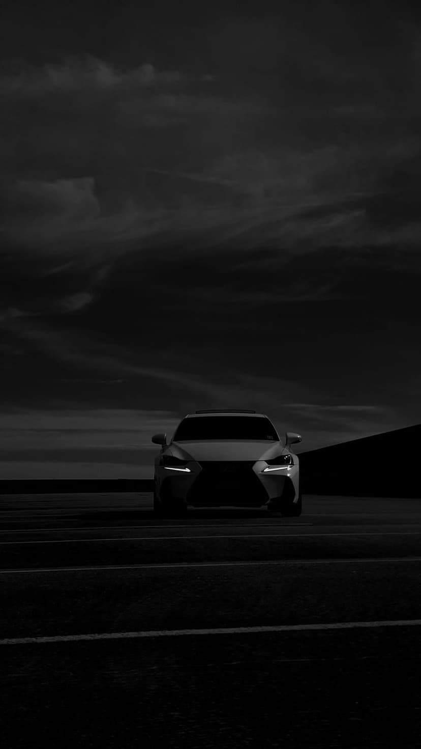 Lexus IS sedan with glowing headlights on a dark asphalt road under a cloudy sky in a monochromatic setting.