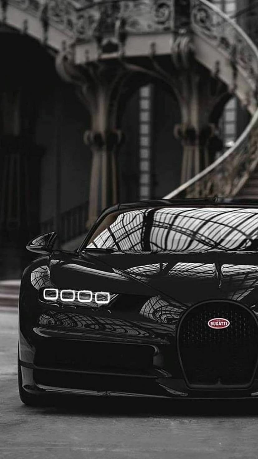 Black Bugatti Chiron parked in a dim grand hall near an ornate marble staircase with glowing LED headlights.