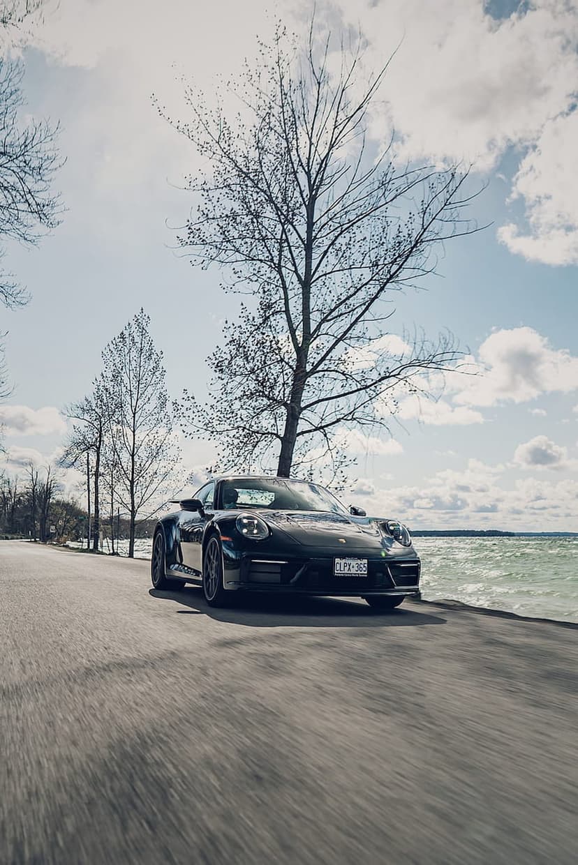 Black Porsche 911 sports car drives along a scenic ocean highway with motion blur and bare trees.