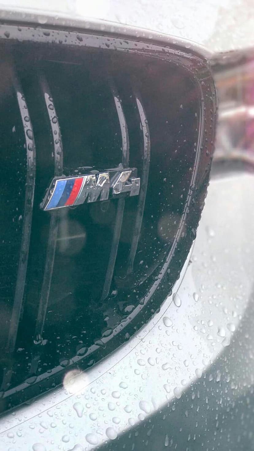 BMW M4 badge on a black honeycomb grille covered in tiny transparent water droplets during a rainy day.