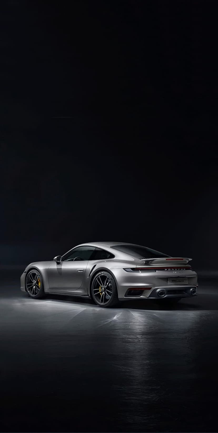 Silver Porsche 911 Turbo S rear view in a dark studio with dramatic lighting and a polished floor reflection.