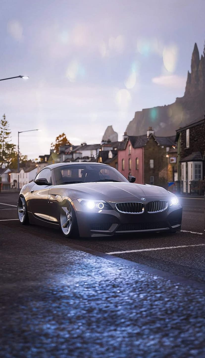 Dark gray BMW Z4 with glowing headlights parked on a wet street at dusk under a moody overcast sky with bokeh lights.