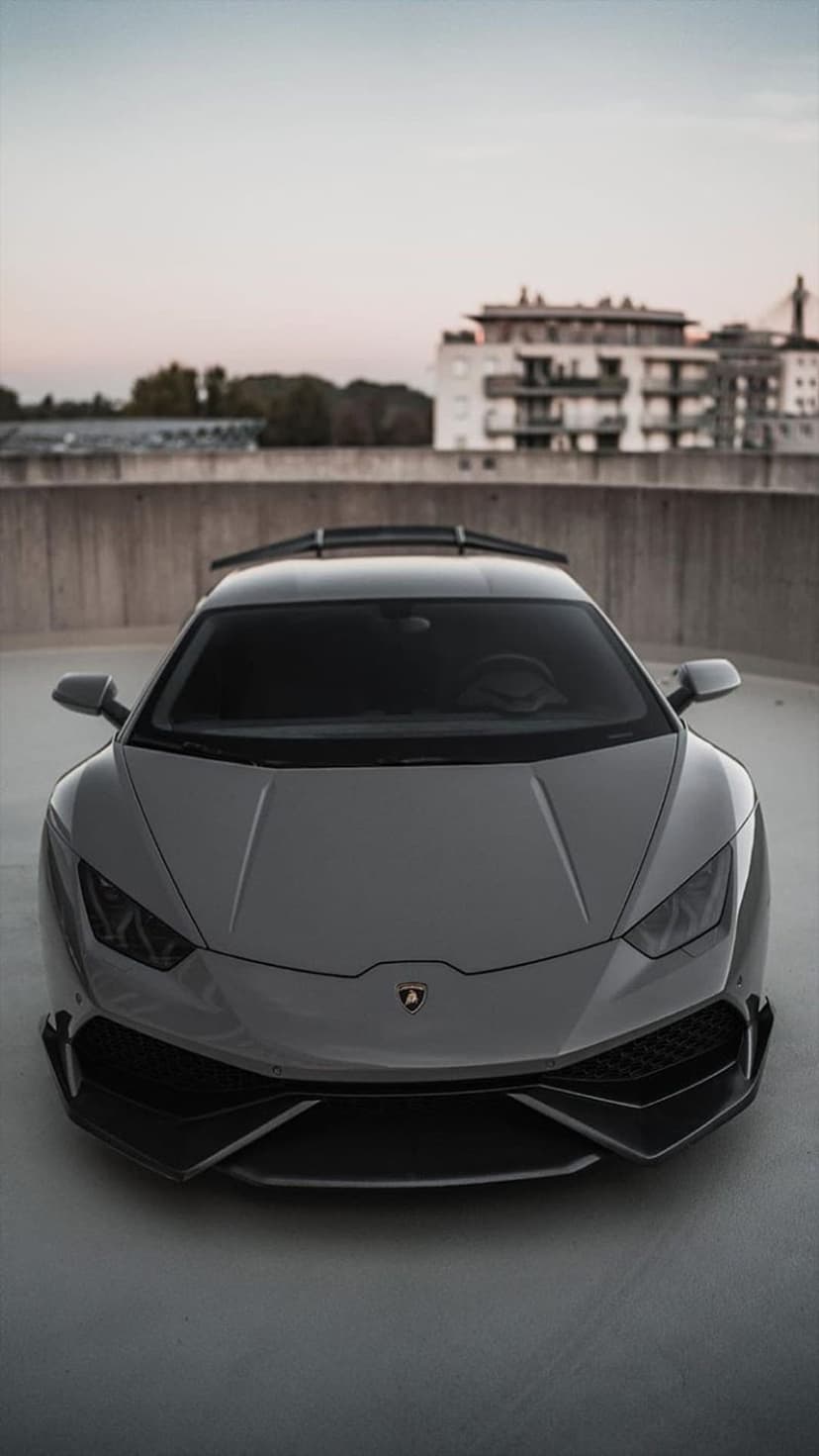 Gray Lamborghini Huracan parked on a concrete rooftop with modern city buildings under a soft twilight sky.