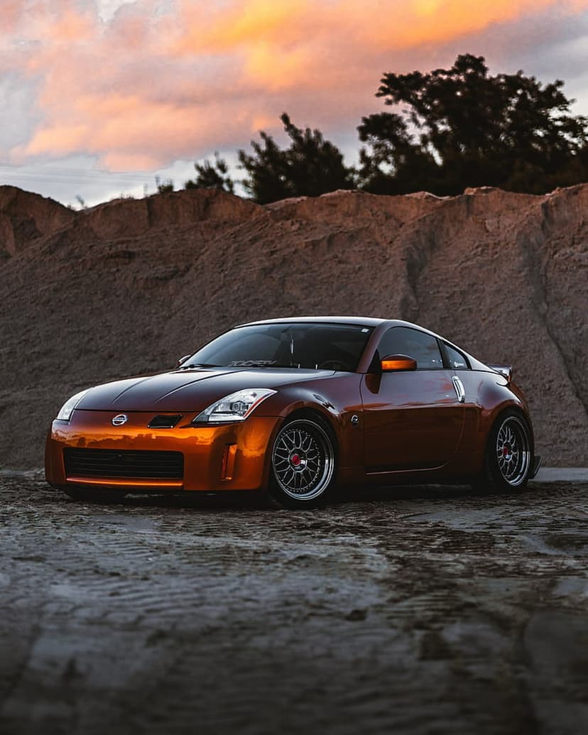 Orange Nissan 350Z parked on a sandy beach under a dramatic sunset sky with custom wheels and a low-angle view.