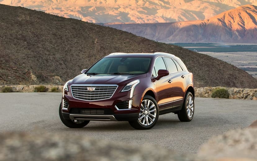Maroon Cadillac XT5 SUV on a paved mountain road with desert peaks and a valley at sunset in the background.
