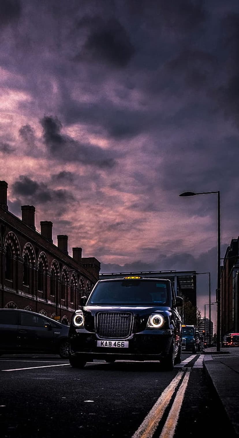 Black London taxi cab with glowing headlights driving on a city street under a dramatic purple and grey dusk sky.