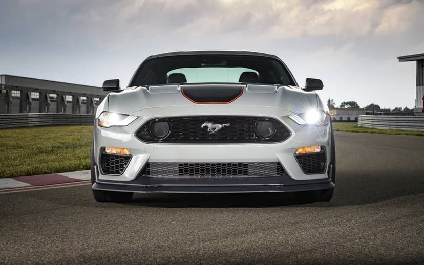 White Ford Mustang Mach 1 with headlights on facing forward on an asphalt racetrack under an overcast sky.