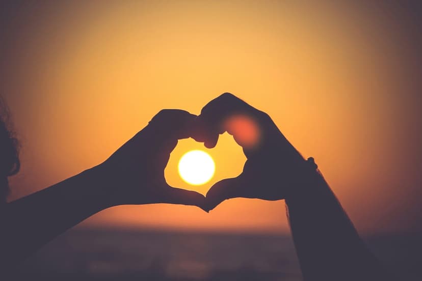 Hands forming a heart shape silhouette against a golden sun over the ocean during a romantic summer sunset.