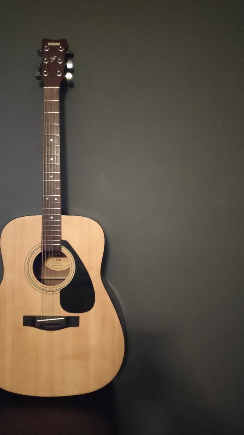 Acoustic guitar with a light wood finish and black pickguard rests against a dark gray wall with subtle shadows.