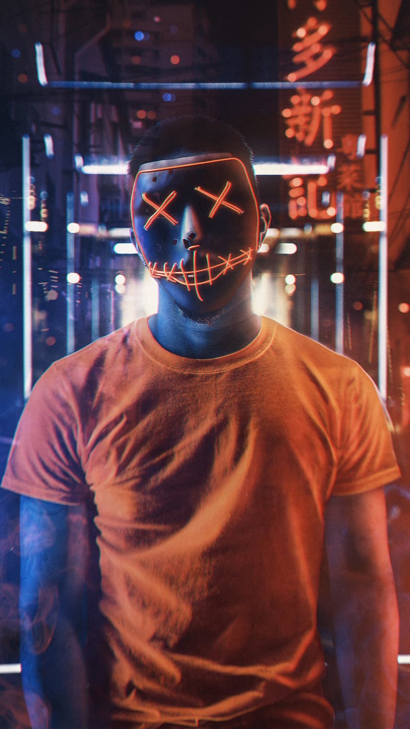 Person wearing a black mask with glowing orange X eyes and a stitched smile against a blurry city at night.