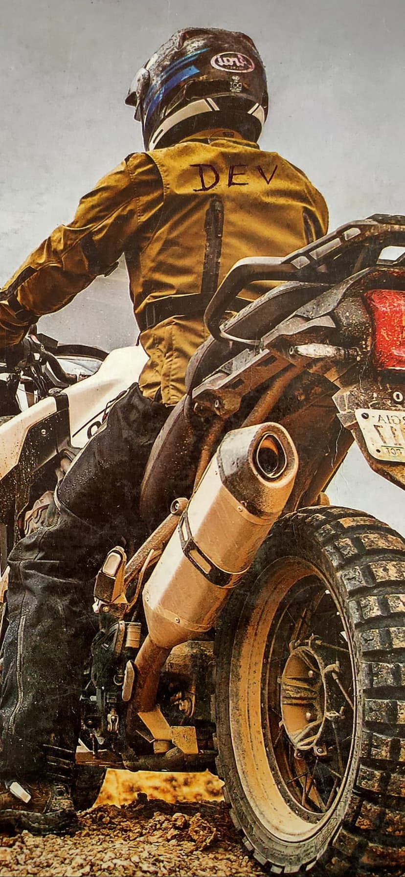 Dirt bike rider wearing a yellow DEV jacket and helmet rides through mud, focusing on the rear tire and exhaust.