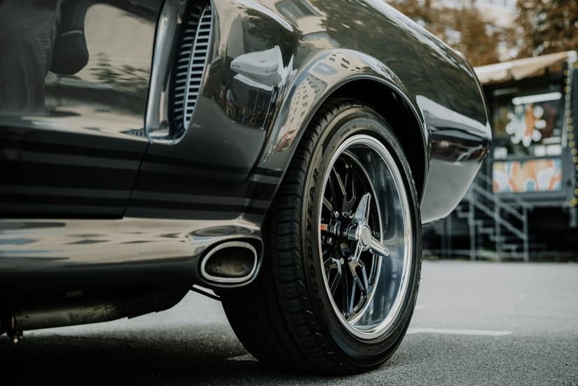 Chrome rim of a dark muscle car with a visible exhaust pipe and tire tread against a blurred urban background.