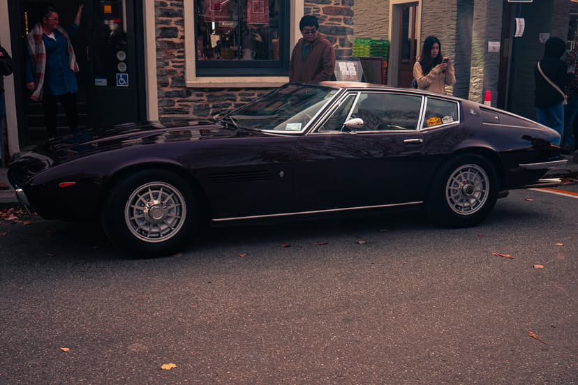 Dark red vintage sports car with wire wheels and chrome trim parked on asphalt near a brick building.