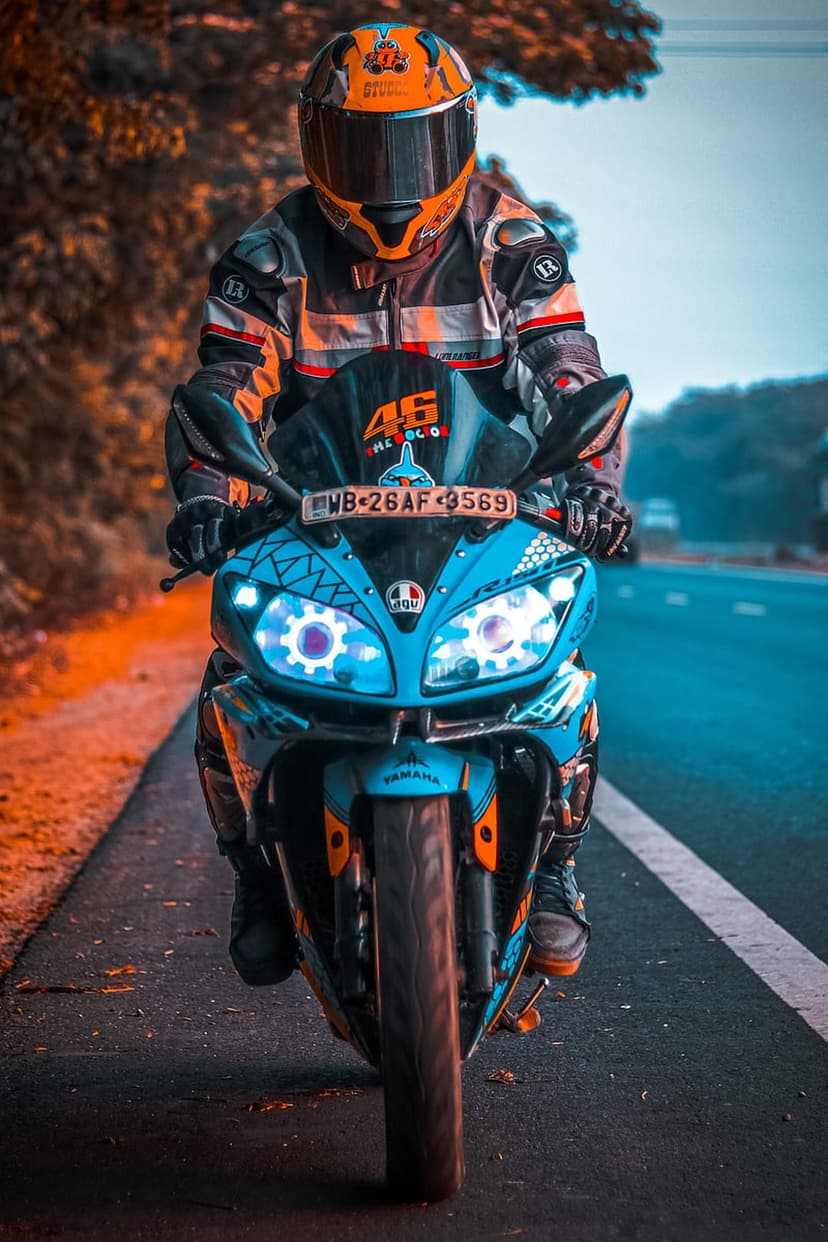 Motorcyclist in orange gear rides a blue Yamaha bike with glowing headlights on a scenic asphalt road.