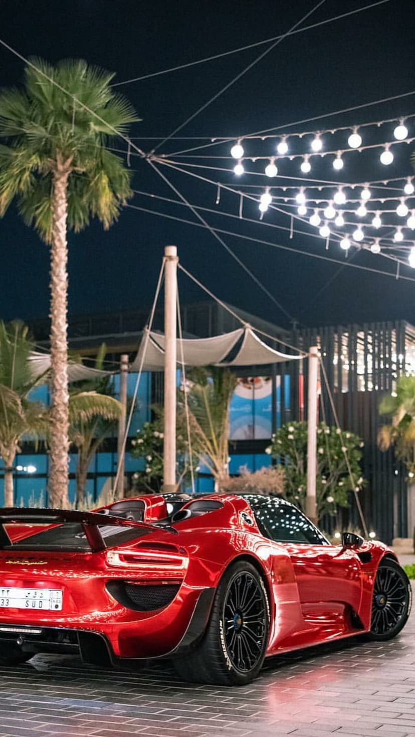 Red sports car parked under glowing string lights and palm trees at night, highlighting sleek vehicle curves.