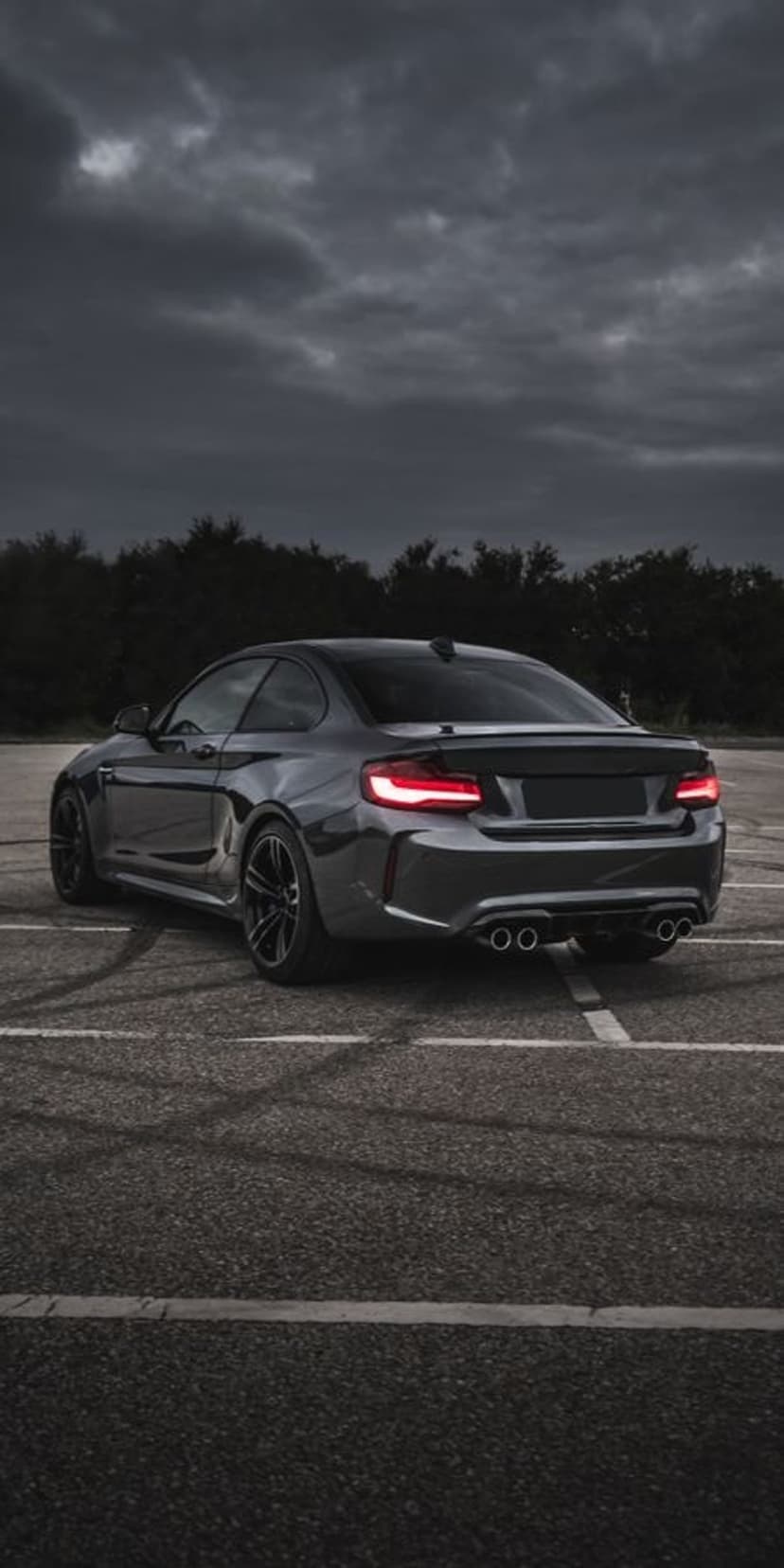 BMW M2 rear view with glowing red taillights and white tire smoke on a dark asphalt road under cloudy skies.