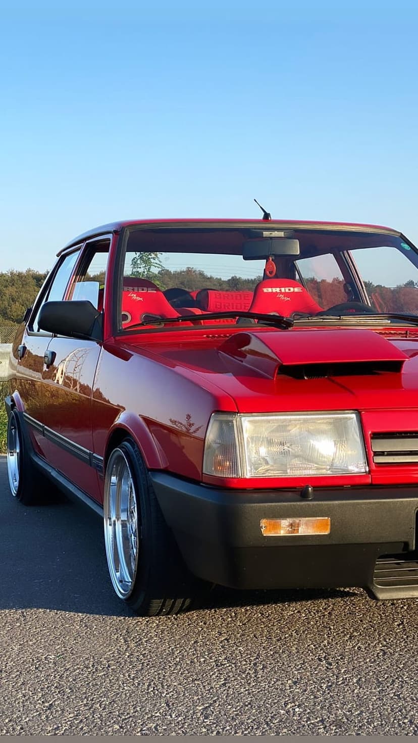 Red modified sedan with racing seats and a hood scoop parked under a clear blue sky on a paved street surface.