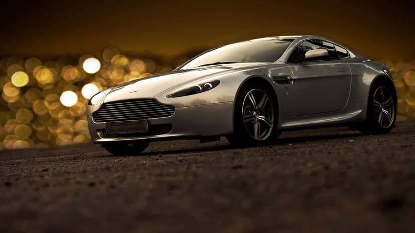 Silver Aston Martin sports car on dark ground with warm golden bokeh lights in the blurred background.