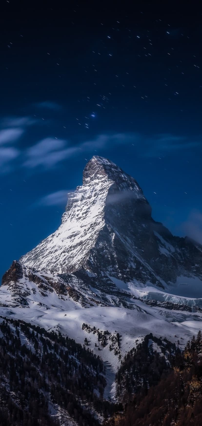 The Matterhorn peak stands under a starry night sky with wispy clouds and snow-covered slopes in Zermatt.