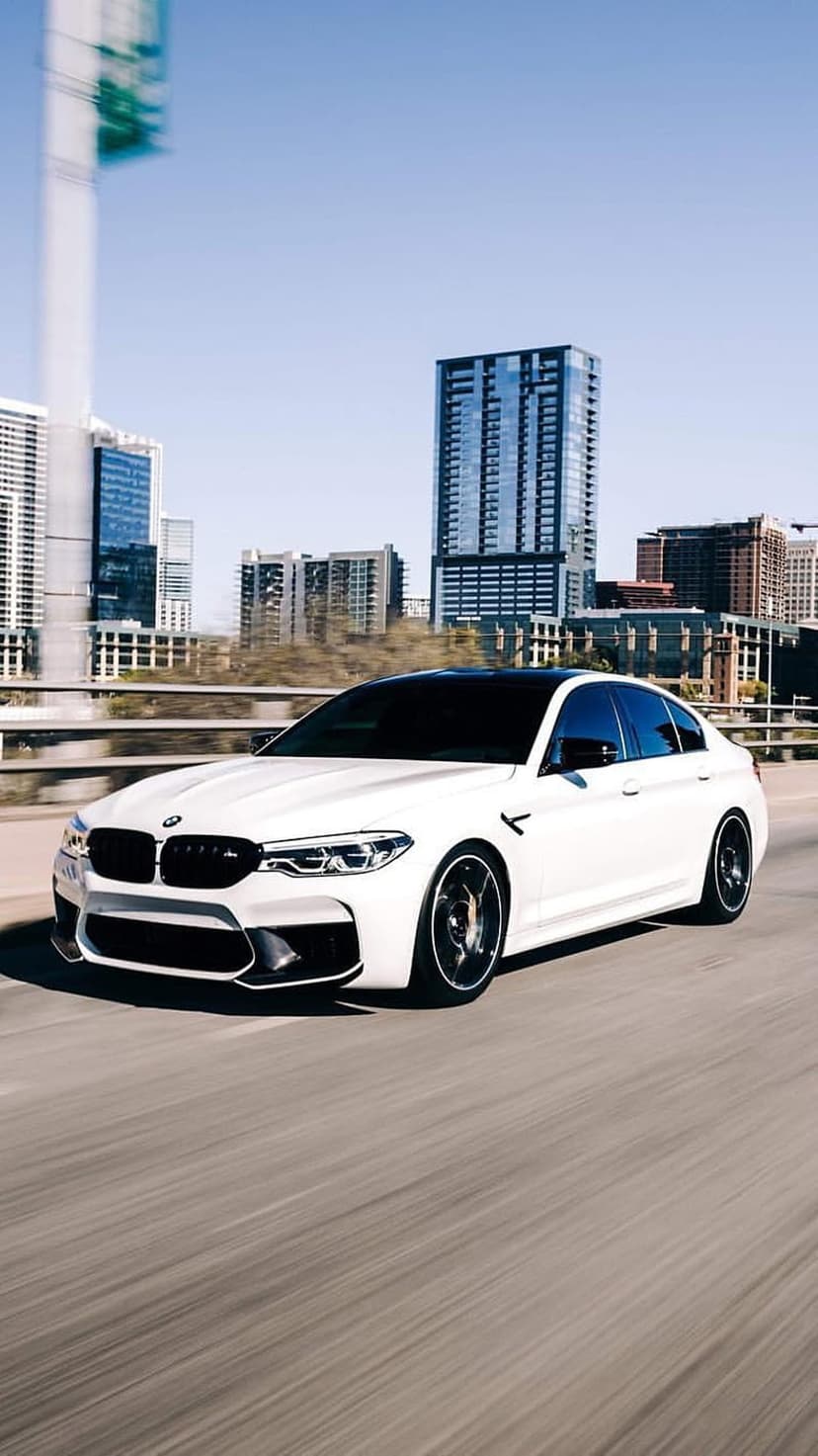 White BMW M5 sedan driving fast on a highway with motion blur and a distant city skyline under daylight.