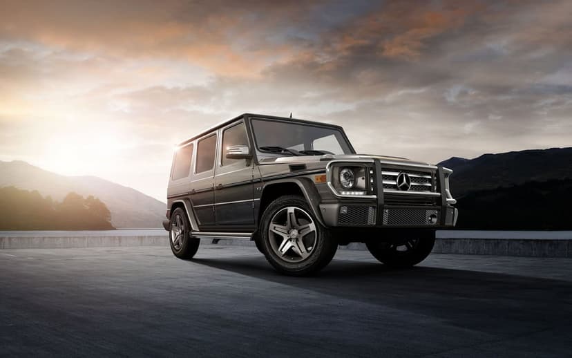 Metallic grey Mercedes-Benz G-Class SUV parked on a paved lot at sunset with distant mountains and a lake.