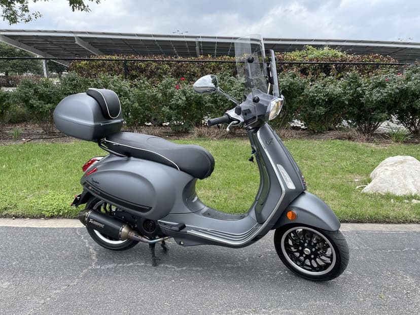 Matte grey Vespa Sprint scooter with Akrapovic exhaust and top box parked on asphalt against green bushes.