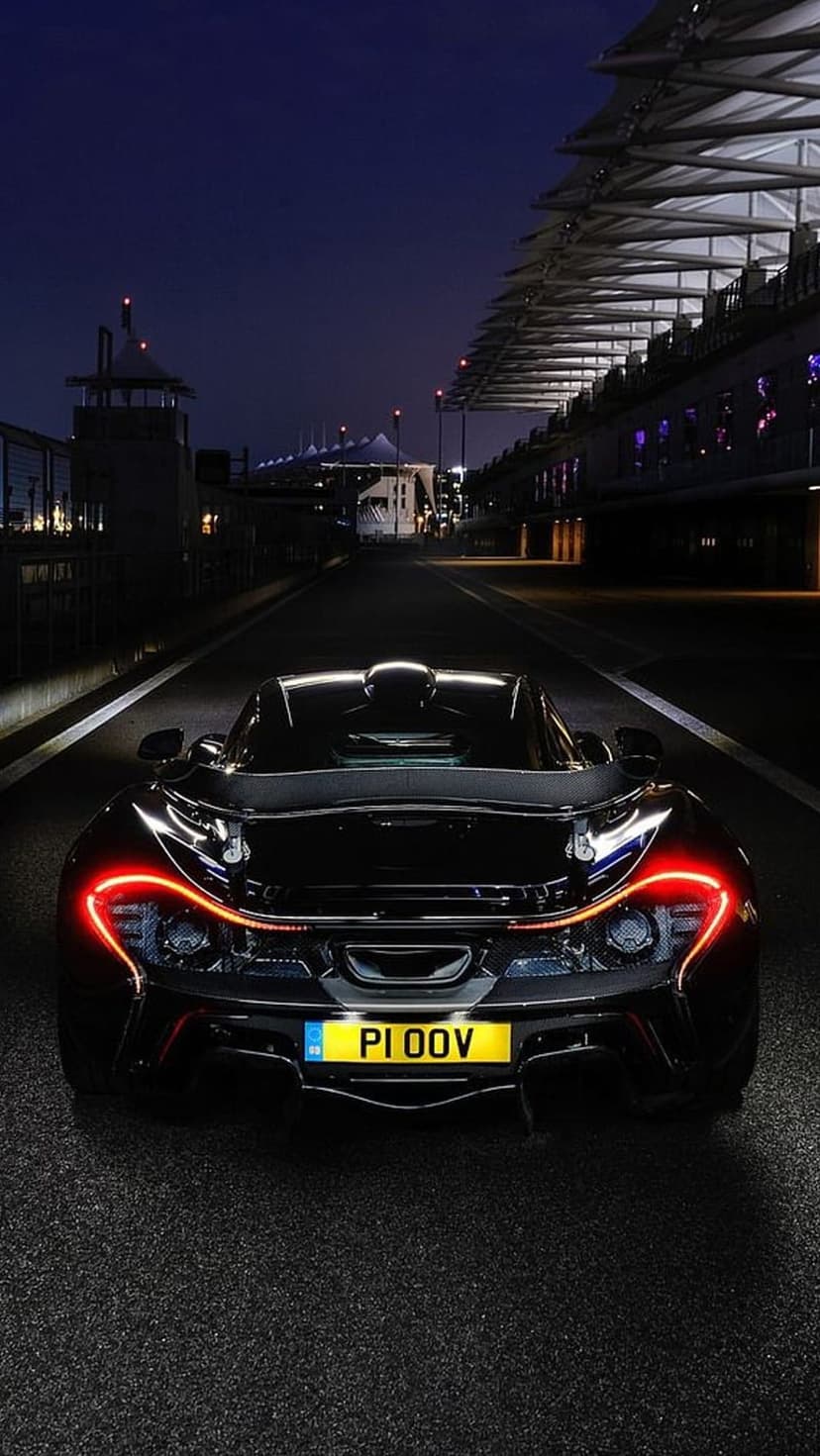 McLaren P1 hypercar with glowing red taillights parked on an asphalt race track under bright stadium lights.