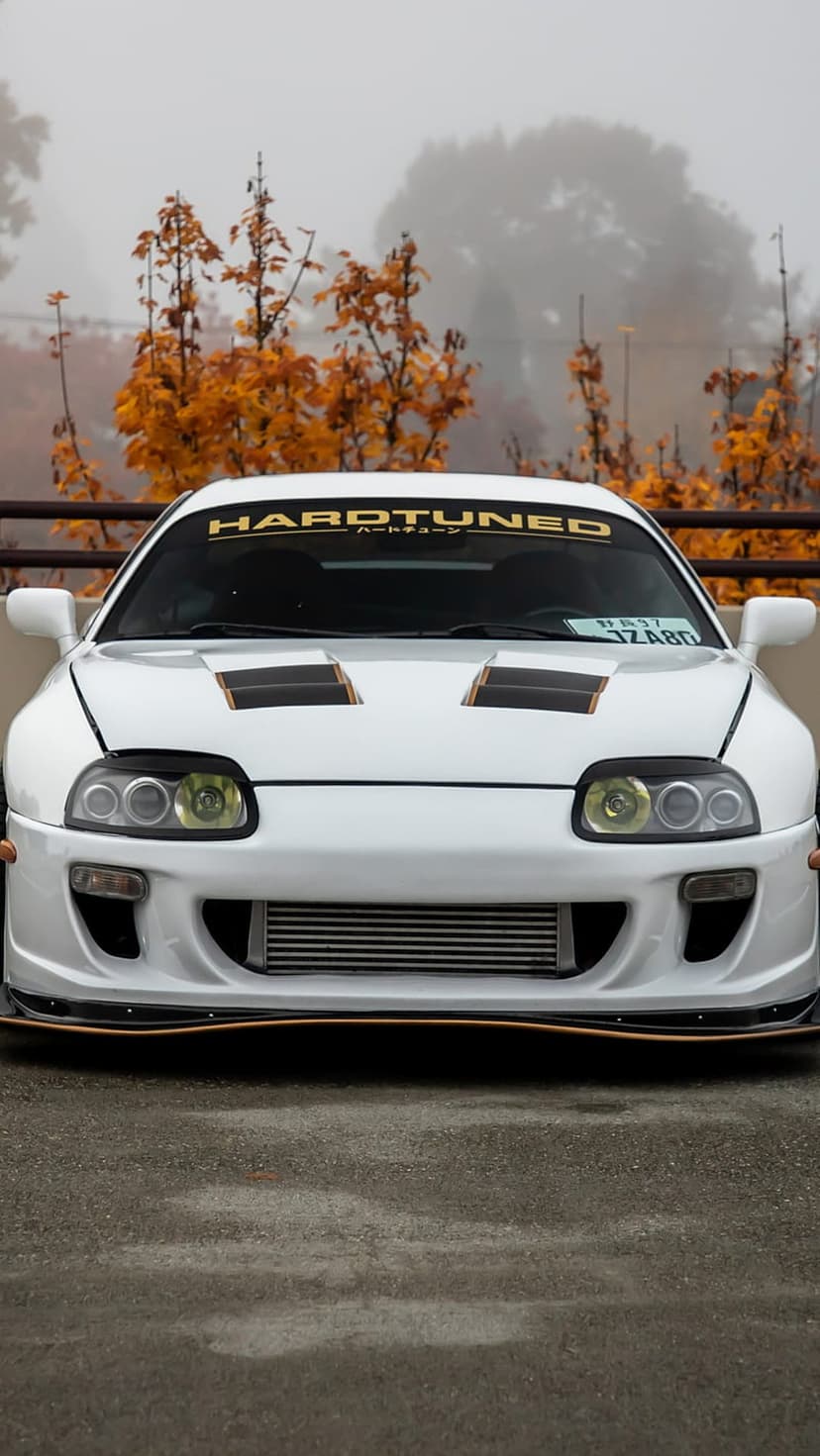 White Toyota Supra MK4 with a windshield decal parked in front of foggy orange autumn trees and golden foliage.