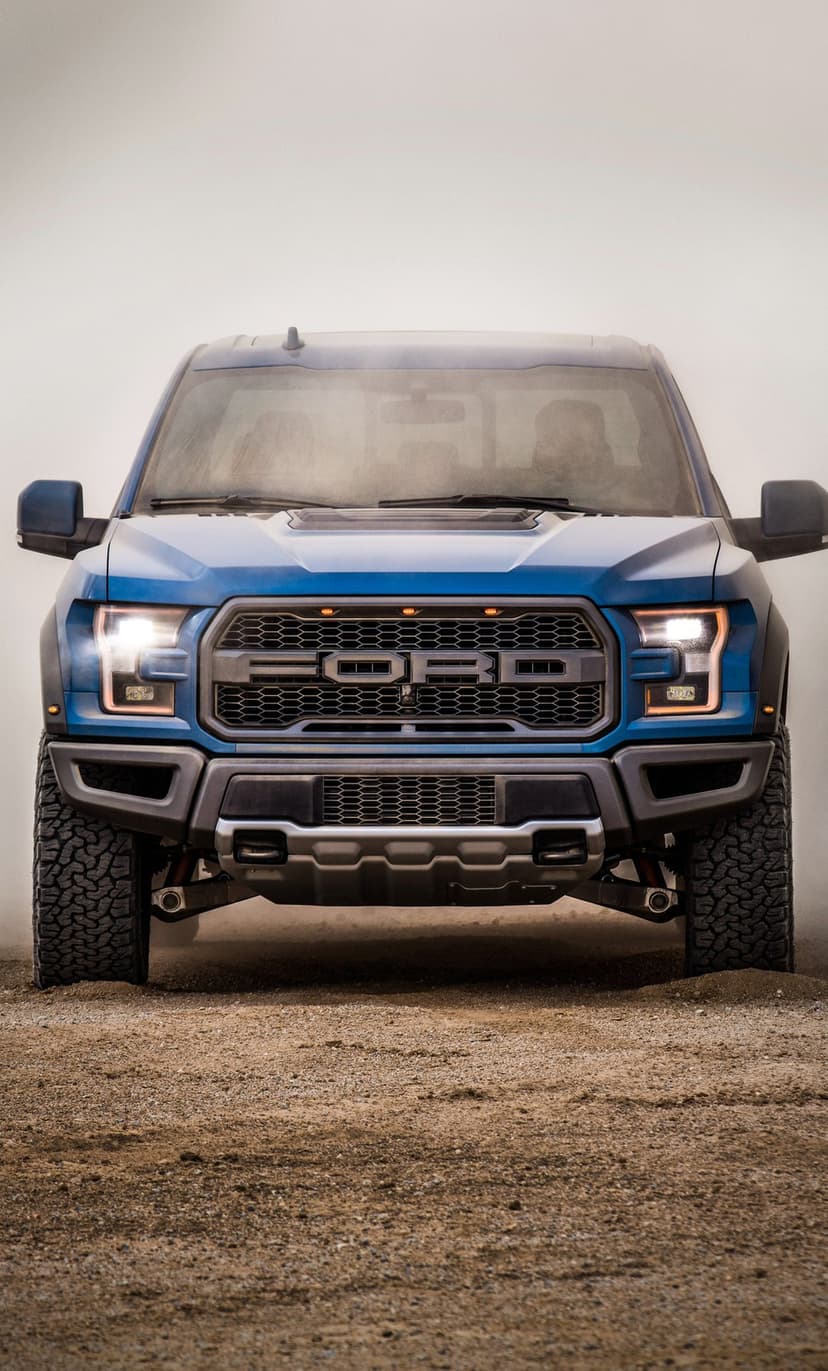 Front view of a blue Ford F-150 Raptor truck with headlights on in a dusty desert environment.