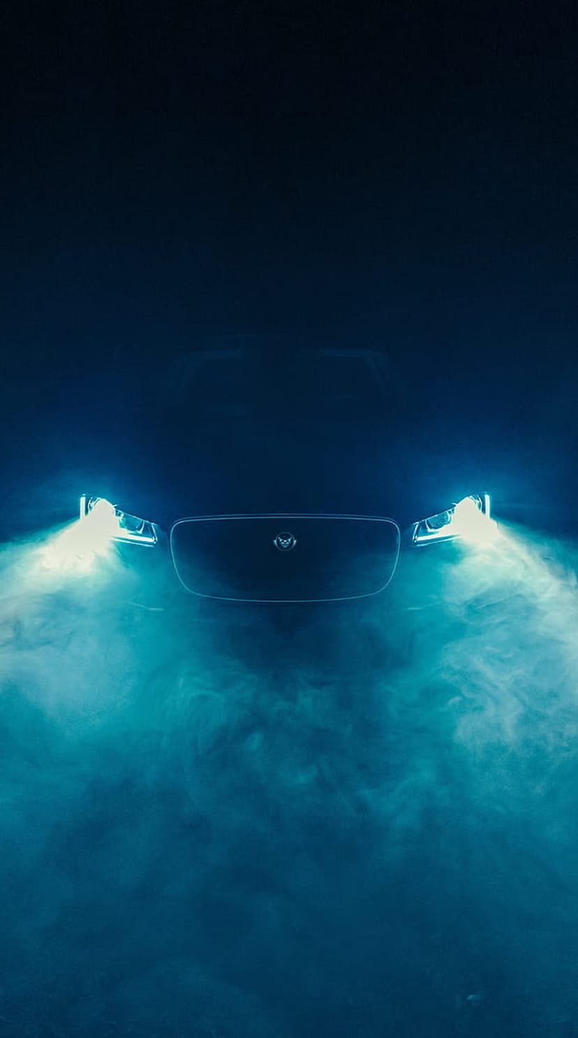 Jaguar car front grille and illuminated headlights emerge from thick blue mist in a vertical night scene.