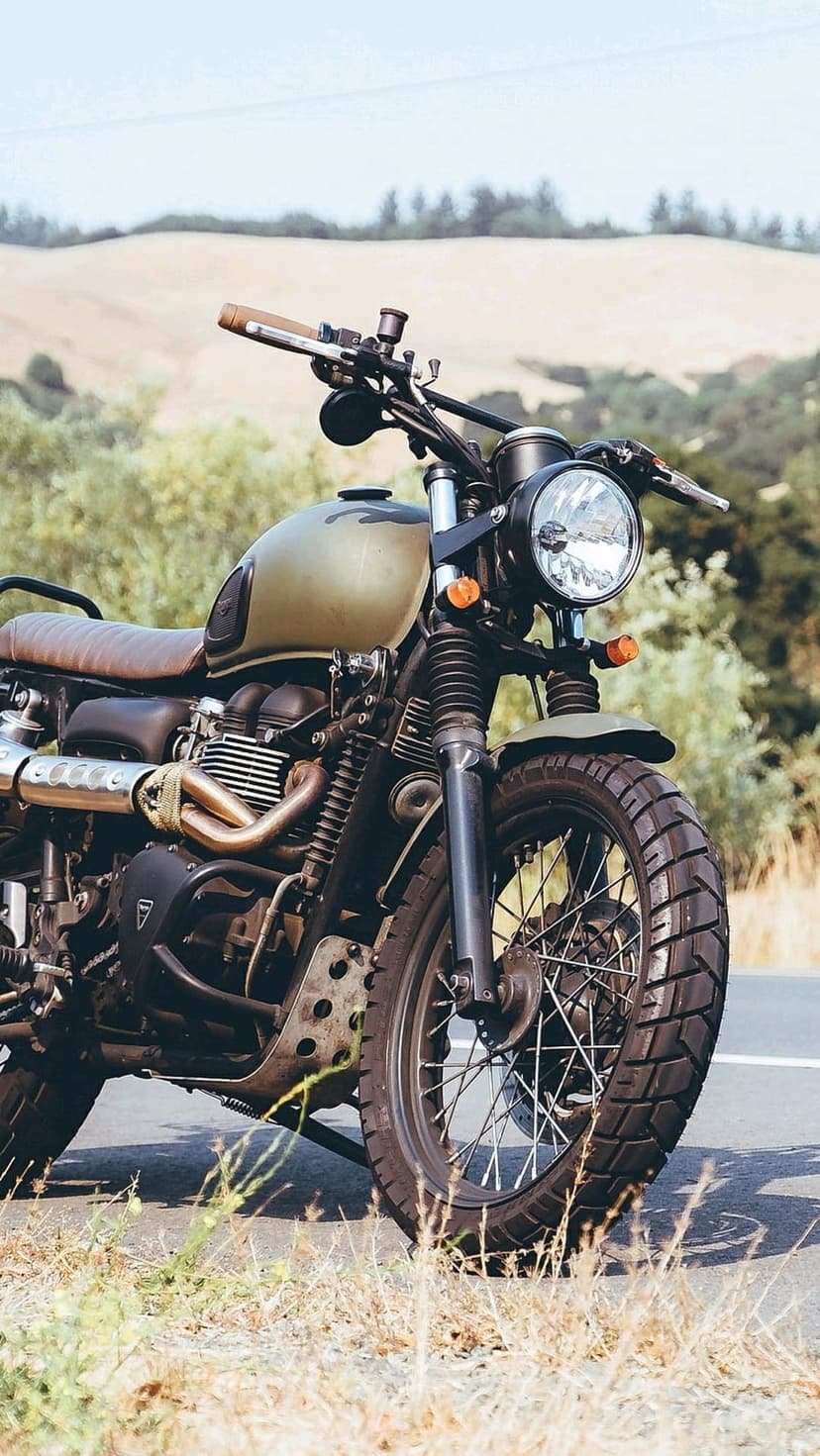 Olive green scrambler motorcycle with a brown leather seat parked on a roadside against golden rolling hills.
