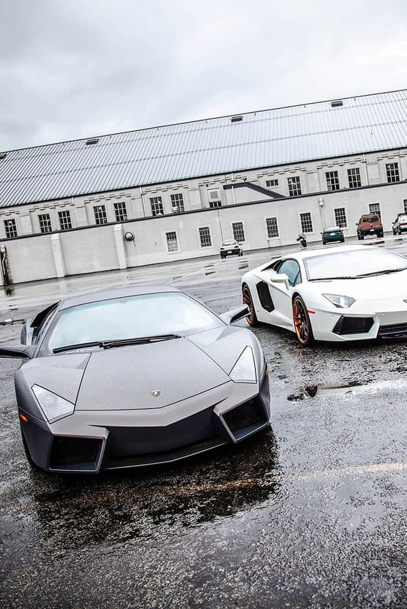 Matte grey Reventon and white Aventador with orange wheels on wet asphalt before a modern white building.