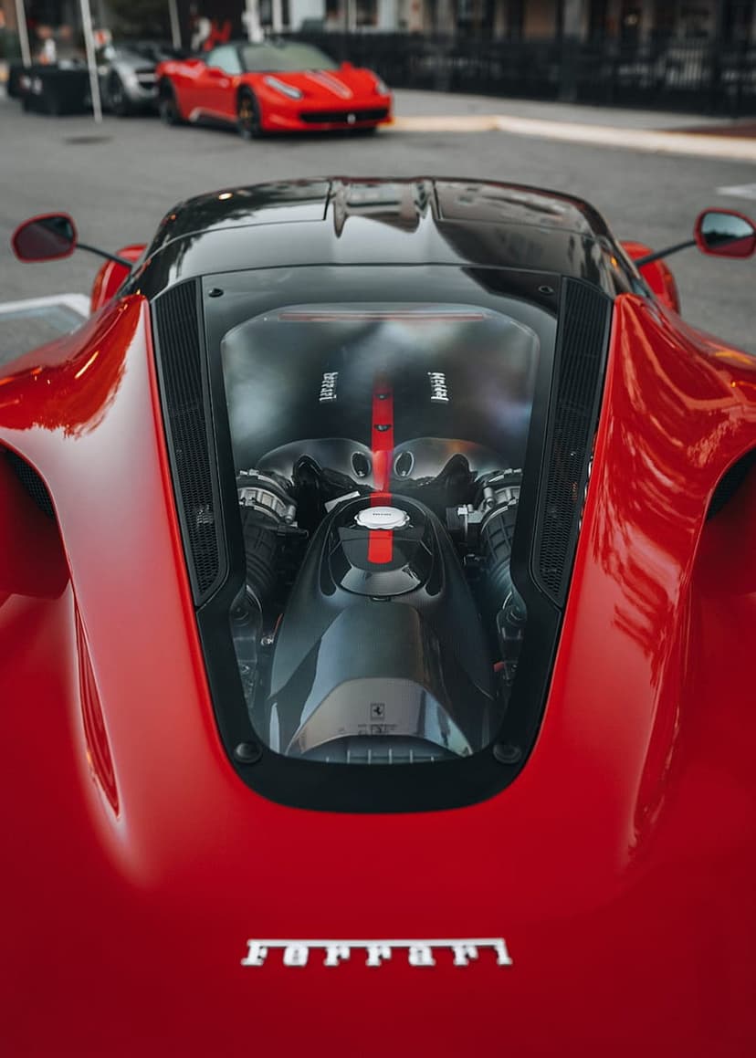 Ferrari V12 engine block with a red stripe and metallic parts, featuring blurred sports cars in the background.