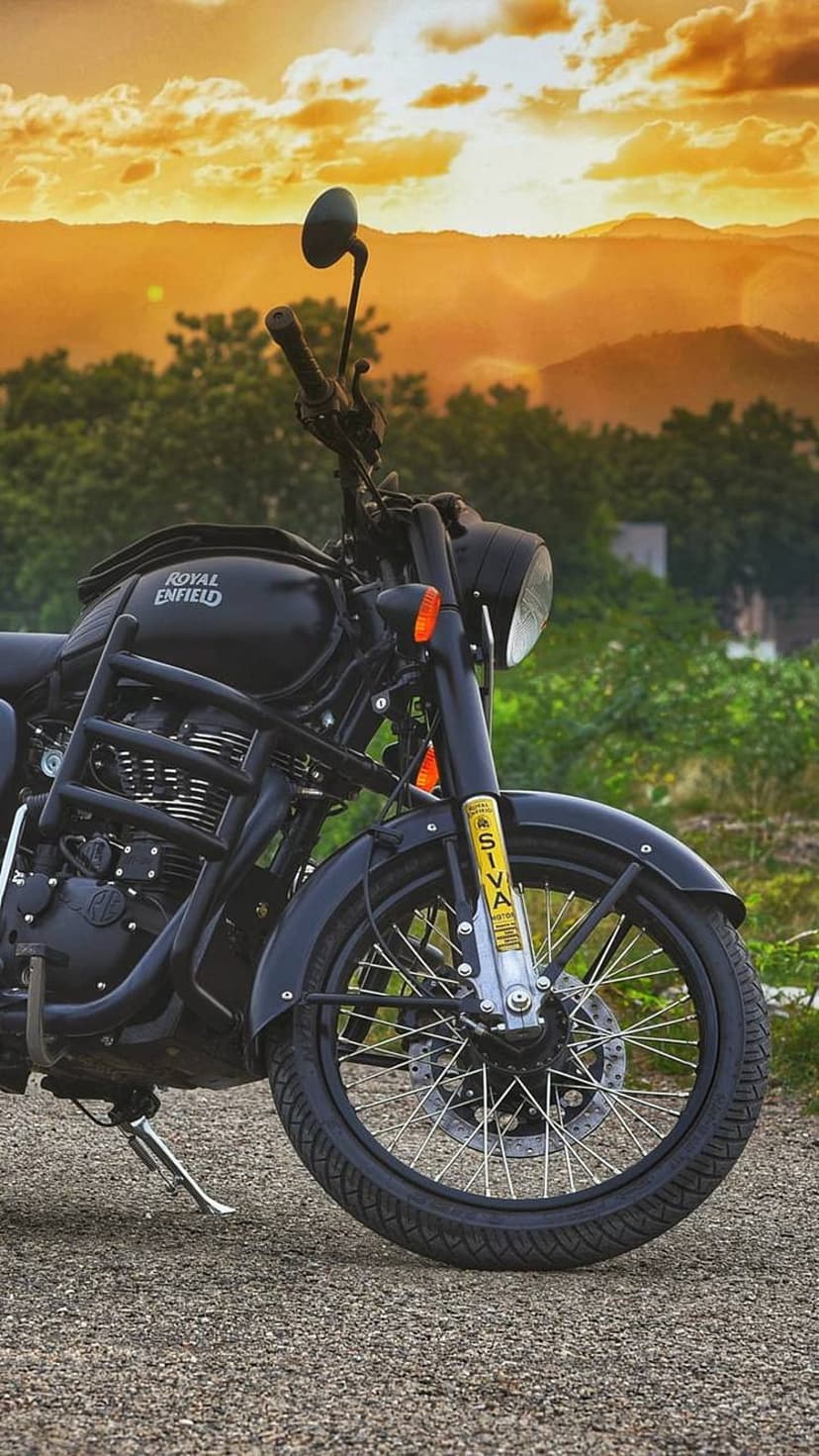Black Royal Enfield motorcycle on a gravel path with rolling green hills under a golden sunset sky.