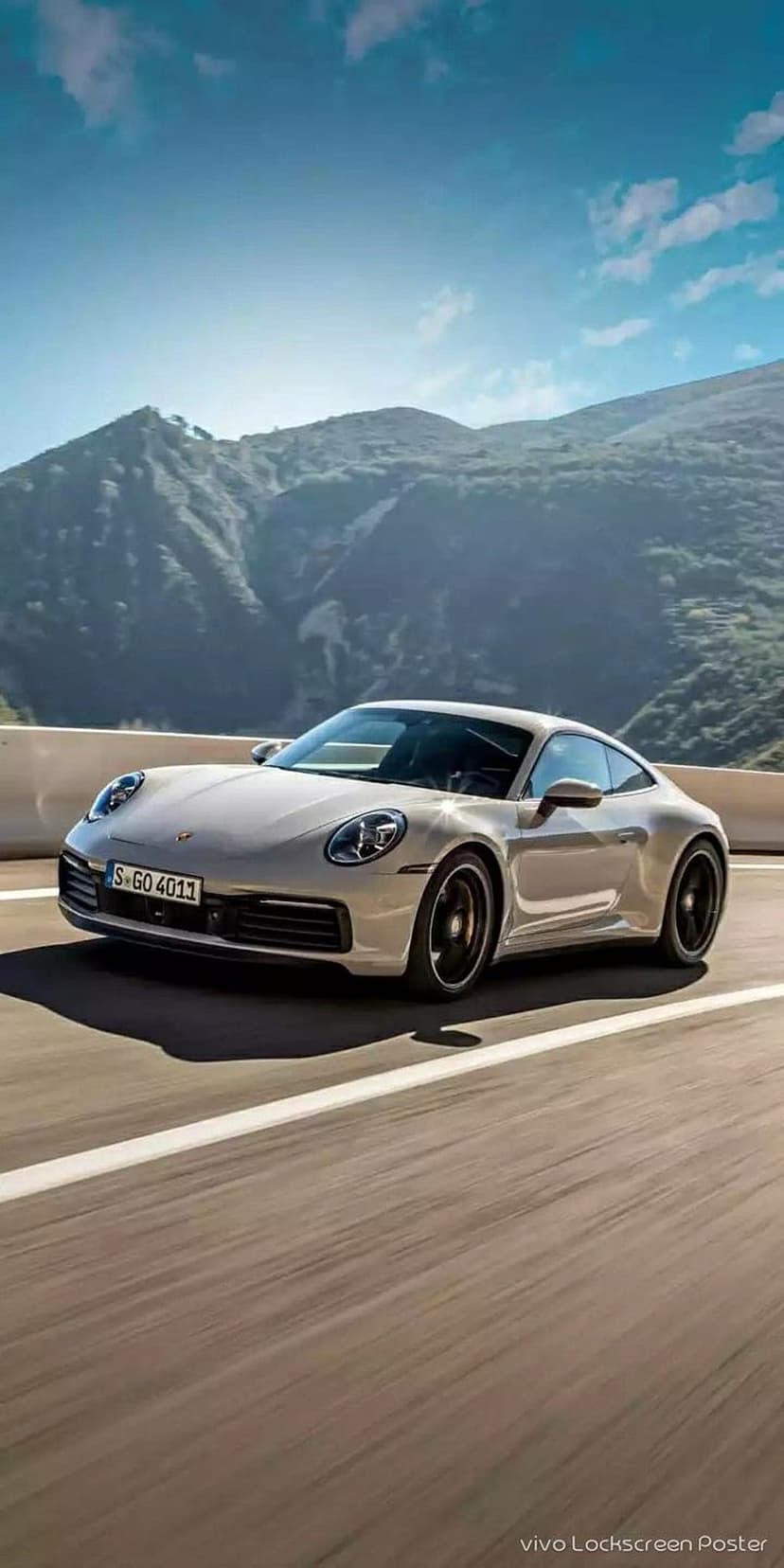 Silver Porsche 911 Carrera driving on a winding mountain road under a clear blue sky with rugged peaks behind it.