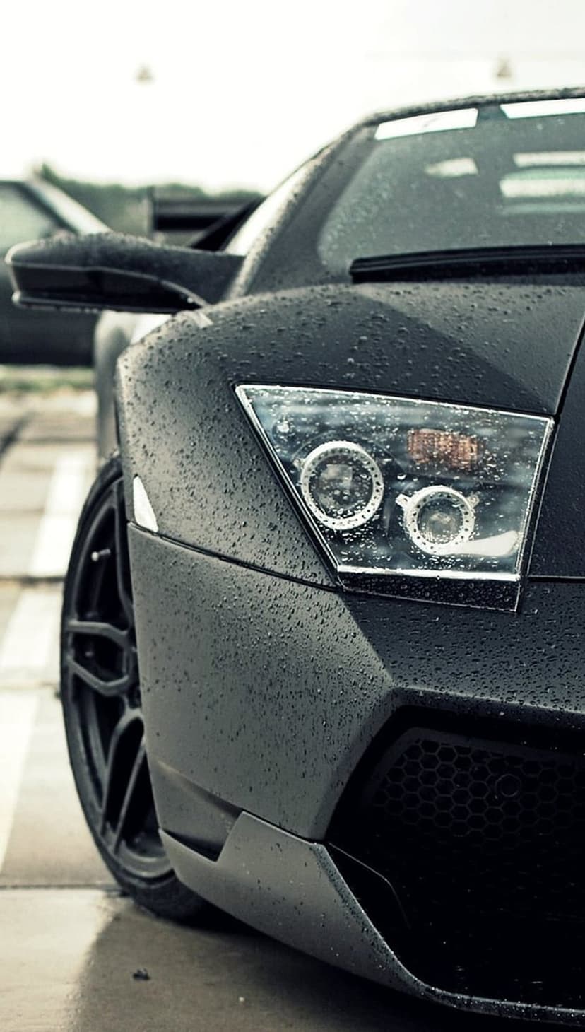 Black Lamborghini headlight and front wheel covered in water droplets under low light for mobile display.