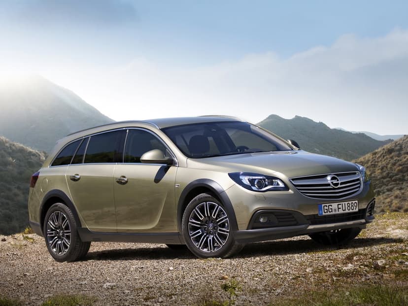 Gold Opel Insignia Country Tourer wagon parked on a gravel mountain road under a bright sunny sky with lens flare.