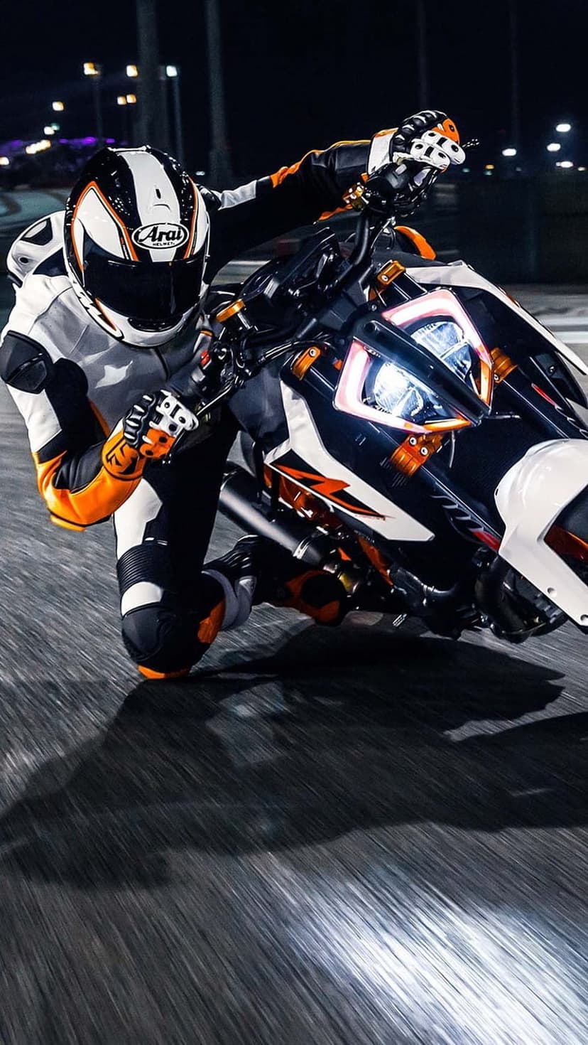 Motorcyclist in white and orange racing leathers leans a bike into a dark turn on a glowing asphalt track.