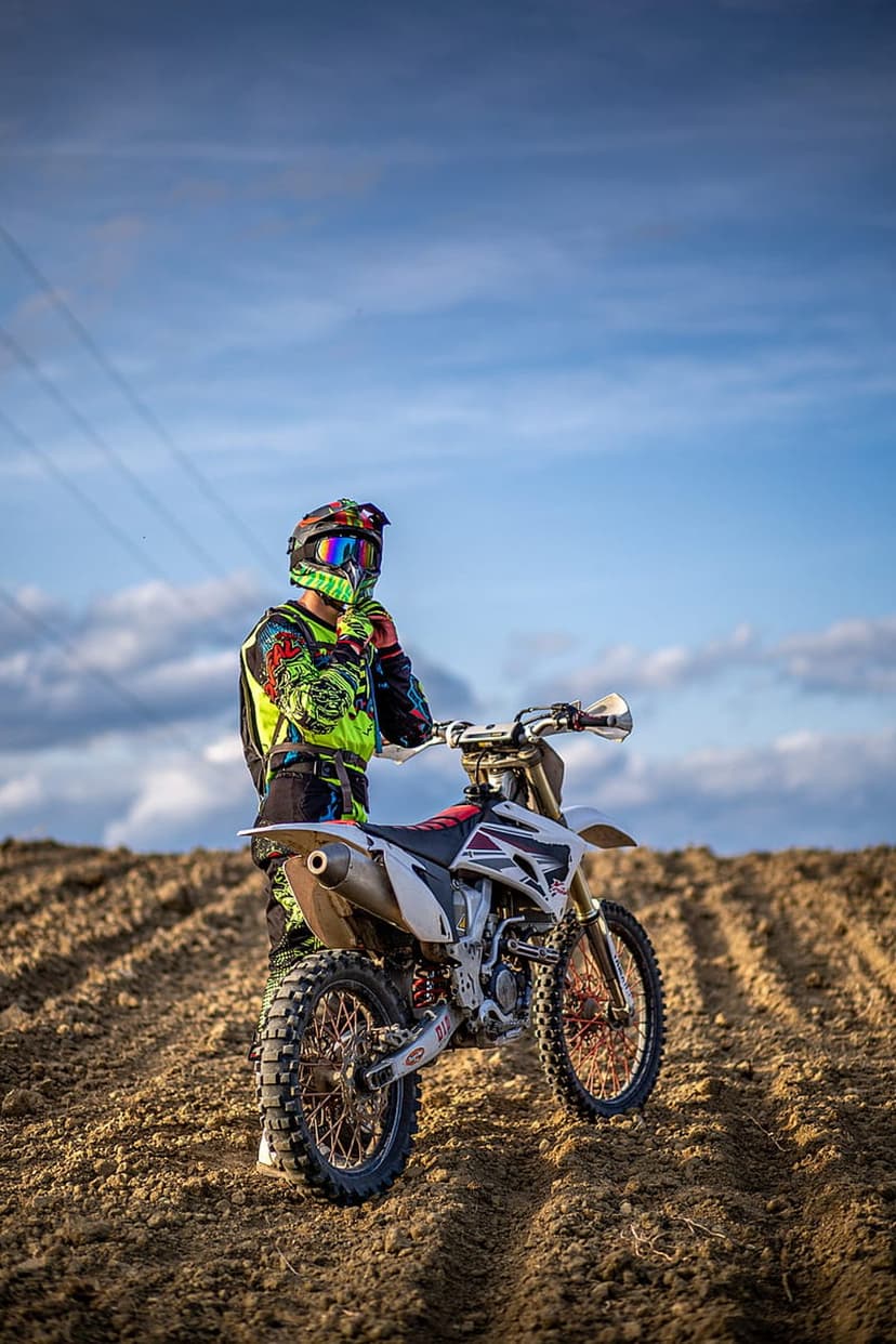 Motocross rider in a colorful racing suit stands next to a white dirt bike in a rural field under a blue sky.